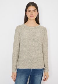 Femme aux longs cheveux foncés portant un pull en maille gris clair et un jean bleu, debout devant un fond blanc uni.
