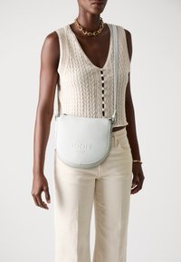 Bela usnjena torba čez ramo z teksturirano površino, vtisnjenim logotipom in nastavljivim trakom. Model nosi pleteno majico in bež hlače.