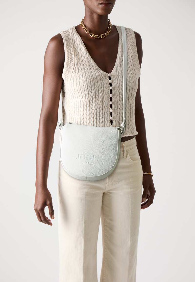 Bela usnjena torba čez ramo z teksturirano površino, vtisnjenim logotipom in nastavljivim trakom. Model nosi pleteno majico in bež hlače.