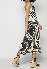 Femme portant une robe midi sans manches à motifs noir et blanc et des sandales compensées beiges avec des brides noires à la cheville, marchant vers la gauche.