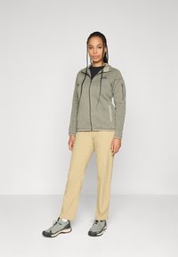 Woman with long braids standing, wearing grey zip-up fleece jacket, beige pants, and grey hiking shoes against a plain white background.