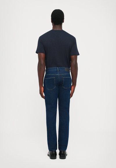Dark blue jeans with a straight leg design, standard five-pocket style, and a leather patch at the waistband. Worn with a black T-shirt.