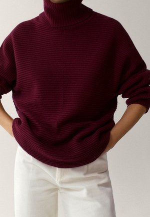 Personne portant un pull à col roulé côtelé bordeaux avec un pantalon blanc, les mains dans les poches, debout devant un fond uni.
