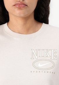 Šviesiai smėlinės spalvos medvilnės marškinėliai su apvaliu iškirpimu. Pabrėžta ryški "NIKE" užrašų ir logotipo kontrastinga spalva, pažymėta "SPORTSWEAR."