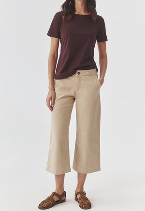 Femme portant un haut marron foncé à manches courtes, un pantalon beige large et court, et des sandales en cuir marron, debout avec une main dans la poche.