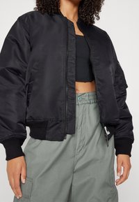 Zwarte bomberjack met een geribbelde zoom en manchetten, gemaakt van gladde, glanzende stof. Heeft een rits aan de voorkant en een zak op de mouw.