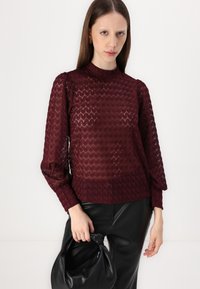 Bordo pleteni top s teksturiranim heksagonalnim uzorkom, visokim izrezom, napuhanim rukavima i rebrastim manžetama, u kombinaciji s crnim kožnim hlačama i torbom.