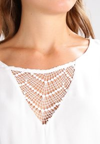Blouse blanche avec des détails en dentelle délicate au niveau de l'encolure, comprenant une découpe triangulaire et un tissu texturé.