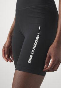 Czarne sportowe szorty o obcisłym kroju, z białym napisem i logo "Swoosh by Nike" z boku, gładki materiał o elastycznej teksturze.