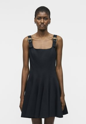 Mujer con vestido negro sin mangas de tirantes anchos con detalles de cadena dorada, posando contra un fondo blanco liso.