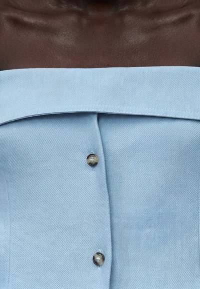 Άτομο με σκουρόχρωμο δέρμα που φοράει ανοιχτό μπλε ρούχο χωρίς ώμους με δύο ορατά στρογγυλά κουμπιά μπροστά.