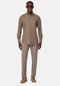 Camisa de manga larga color beige con cuello, combinada con pantalones claros beige y zapatillas de deporte beige. El look se completa con gafas de sol oscuras.