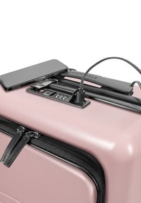 Hartschalenkoffer in sanftem Pink mit schwarzem Reißverschluss, strukturiertem Griff, integriertem Ladeanschluss und einem Smartphone, das oben aufliegt.