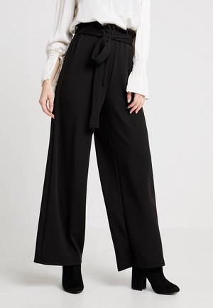 Kvinde iført sorte, højtaljede bukser med vide ben og stofbælte, kombineret med en hvid bluse med lange ærmer og sorte støvler med hæl.