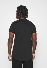 Tricou scurt negru cu mânecă scurtă, cu tiv drept și mâneci răsucite, fabricat dintr-un material moale. Include o etichetă mică cu logo pe lateral.
