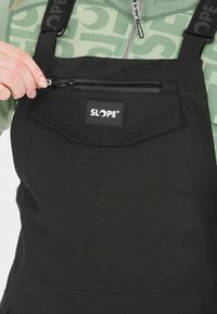 Schwarze wasserdichte Latzhose mit verstellbaren Trägern, ausgestattet mit einer Reißverschlusstasche auf der Brust und einem Logo-Patch. Glatter Stoff, minimalistisches Design.