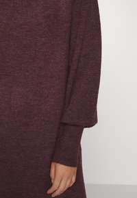 Pull en maille bordeaux à coupe ample, manches larges et poignets côtelés. Texture douce avec de subtils éclats répartis dans le tissu.