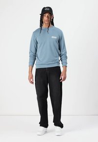Blå genser med lange ermer, hvit logo-patch, kombinert med svarte vidde bukser og hvite sneakers, toppet med en svart caps.