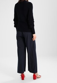 Black knitted sweater with layered ruffle sleeves, paired with navy wide-leg pants featuring a vertical pinstripe pattern, and red block-heeled sandals.