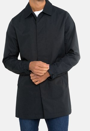 Homme portant un manteau noir boutonné par-dessus une chemise blanche et un jean bleu, les mains jointes à la taille, sur un fond uni.
