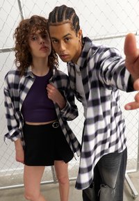 Camicie oversize a scacchi in bianco e nero su due persone; una indossa un top corto viola e l'altra indossa una camicia bianca sotto la camicia a scacchi.
