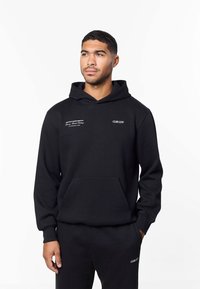 Zwarte hoodie met een voorvak, voorzien van witte tekstbranding op de borst en linkermouw. Gemaakt van zachte stof met een losse pasvorm.