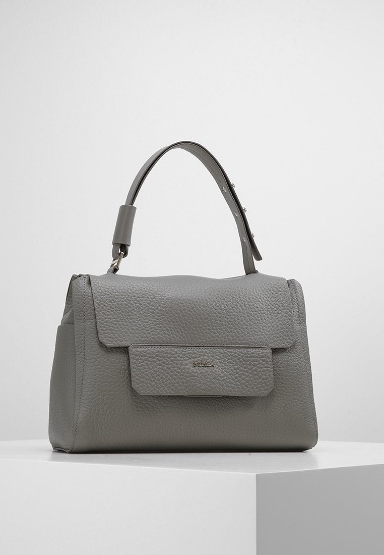 Sac à main en cuir gris texturé avec rabat de fermeture et poignée supérieure réglable sur un piédestal blanc contre un fond uni.