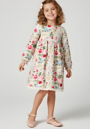 Młoda dziewczyna uśmiecha się, ubrana w kremową sukienkę z długim rękawem w różowo-czerwono-niebieskie kwiatowe wzory, z spinką do włosów w kształcie stokrotki oraz różowe buty typu Mary Jane.