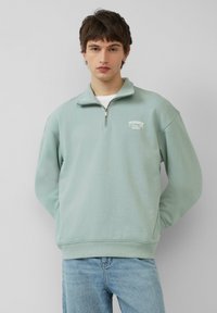 Light green zip-up sweatshirt with a high collar, featuring a small white graphic on the chest and ribbed cuffs. Smooth cotton texture.