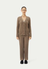 Brauner gerippter Strickcardigan mit V-Ausschnitt und Knopfleiste, kombiniert mit passenden plissierten Hosen. Der Stoff hat eine weiche Textur. Schwarze Schuhe runden den Look ab.