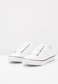 Weiße Leinenschuhe mit niedrigem Schnitt, schwarzer Gummisohle und vorderer Schnürung. Mit Logo an der Seite und rundem Zehenbereich.