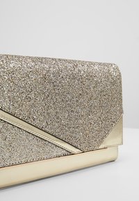 Goldglitzer-Clutch mit metallischem Goldbesatz, dreieckiger Klappe und gesteppten Details vor neutralem Hintergrund.