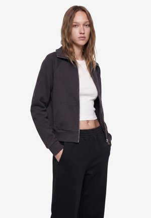 Ung kvinde iført en sort lynlåstrøje, hvid cropped top og sorte bukser med hænderne i lommerne, stående mod en ensfarvet baggrund.