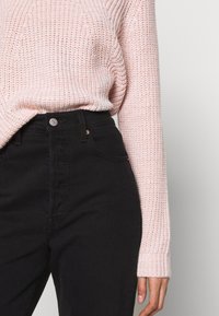 Hellrosa, strukturierter Strickpullover, kombiniert mit hoch geschnittenen schwarzen Jeans mit Knopf- und Reißverschluss.