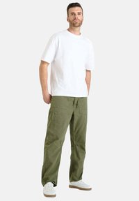 Man in een witte T-shirt met korte mouwen, olijfgroene cargo-broek en witte sneakers, staande met handen ontspannen langs de zijden.