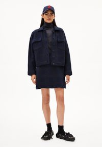 Marinejacke mit zwei Brusttaschen, Knopfverschluss, ergänzt durch einen passenden Rock. Schwarze Schuhe mit Fransen und goldenen Akzenten.