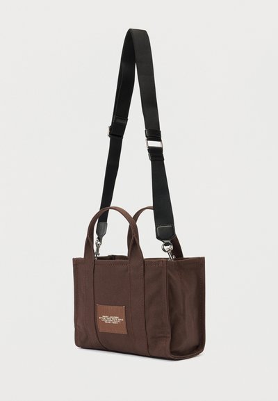 Hnedá obdĺžniková taška s krátkymi rukoväťami a nastaviteľným čiernym ramenným popruhom, s malou prednou nálepkou s textom.