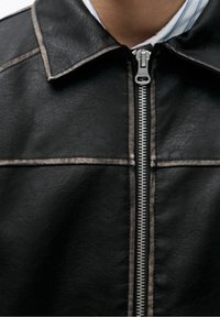 Black leather jacket with a visible silver zipper, pointed collar, and contrasting stitching along the seams. Textured surface detail evident.