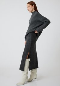 Pull en tricot gris à col haut, associé à une jupe midi gris ajustée avec des fentes latérales et des bottes blanches à bout pointu.