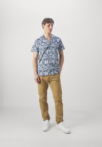Cămașă cu mânecă scurtă de culoare albastră, cu modele geometrice, asortată cu pantaloni bej deschis și adidași albi. Design simplu al gulerului.