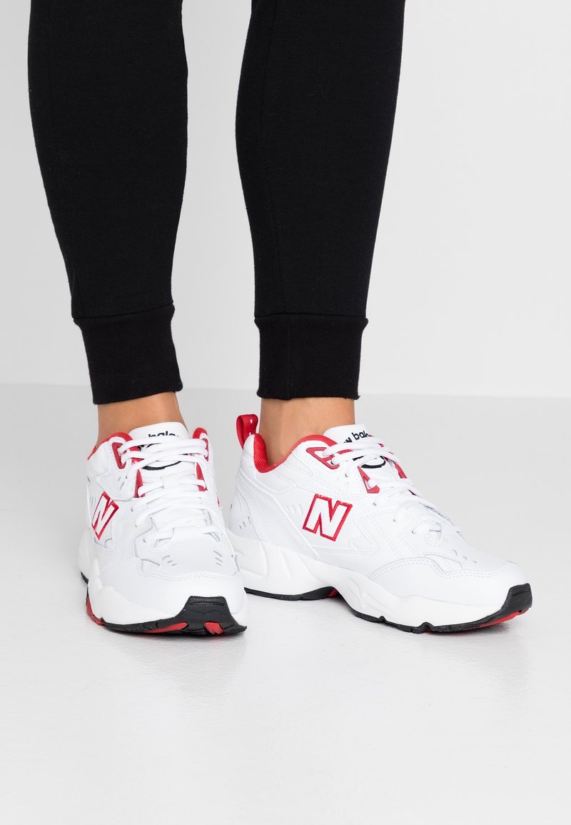 Personne portant des baskets New Balance blanches et rouges avec des lacets blancs et des semelles noires, associées à un pantalon noir ajusté.