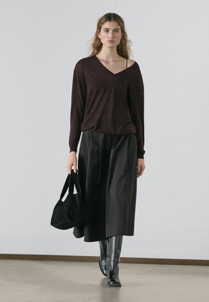 Kvinde iført mørkelilla off-shoulder trøje, sort midi-nederdel, sorte støvler og bærende en sort håndtaske, stående indendørs på neutral gulv.