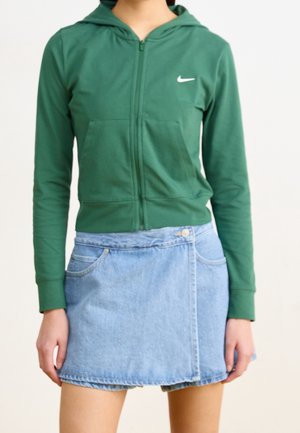 Kobieta w zielonej bluzie Nike na zamek i jasnoniebieskiej dżinsowej mini spódnicy na jednolitym tle.