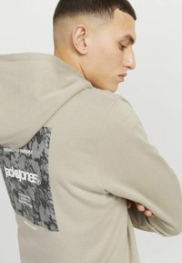 Beige hooded sweatshirt featuring a dark graphic patch on the back with text. The fabric appears soft with a relaxed fit.