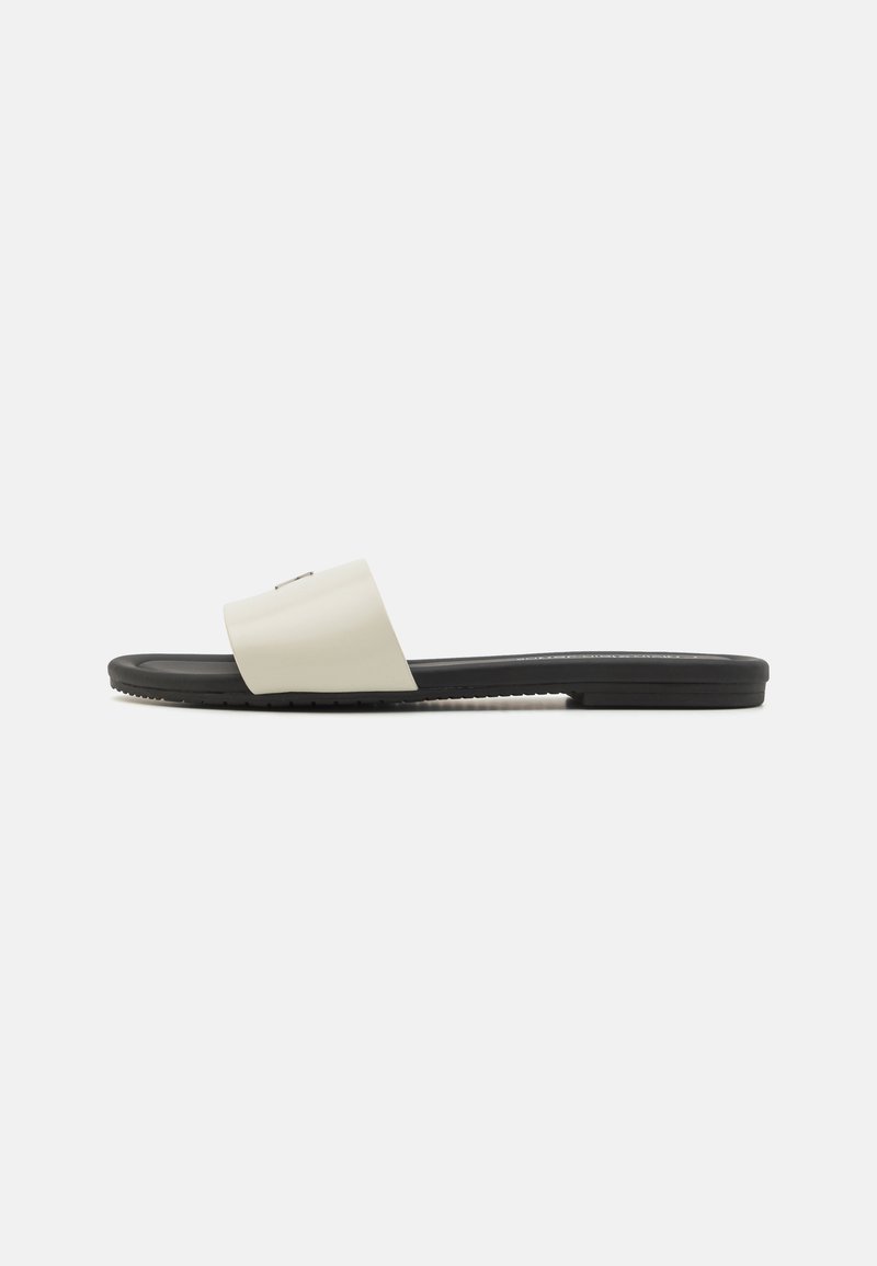 Sandale plate avec une large bride crème et une semelle noire. Fabriquée en matériau lisse avec un design minimaliste et un logo subtil.