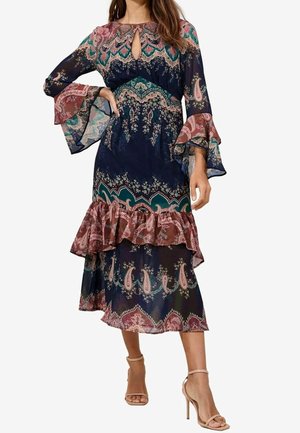 Mujer con vestido midi con patrones de cachemira y flores, mangas acampanadas, detalles de volantes, en azul marino, rosa y teal, combinado con tacones altos beige.