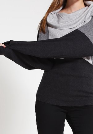 Kobieta ubrana w szaro-czarny sweter z długim rękawem, naciągająca czarny rękaw, w czarnych spodniach i widocznymi długimi brązowymi włosami.