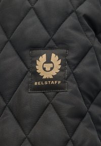Κοντινό πλάνο μαύρου καπιτονέ υφάσματος με μαύρο ετικέτα που φέρει χρυσό φτερωτό έμβλημα και το κείμενο "BELSTAFF".