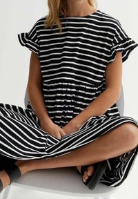 Mustavalkoinen raidallinen mekko, jossa on lyhyet röyhelöhiha, elastinen vyötärö ja aaltoileva hame. Malli istuu tuolissa jalat ristissä.