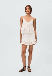 White tiered tank dress with thin straps, textured fabric, ruffled layers, and delicate trim. Paired with black flat sandals.